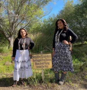photo of two Ohlone sisters standing by a sign that reads "sensitive ecosystem red-legged frog habitat please avoid playing around the pool."